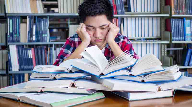 A frustrated pre-med student studying for the MCAT in a library, sitting in front of a pile of open textbooks.
